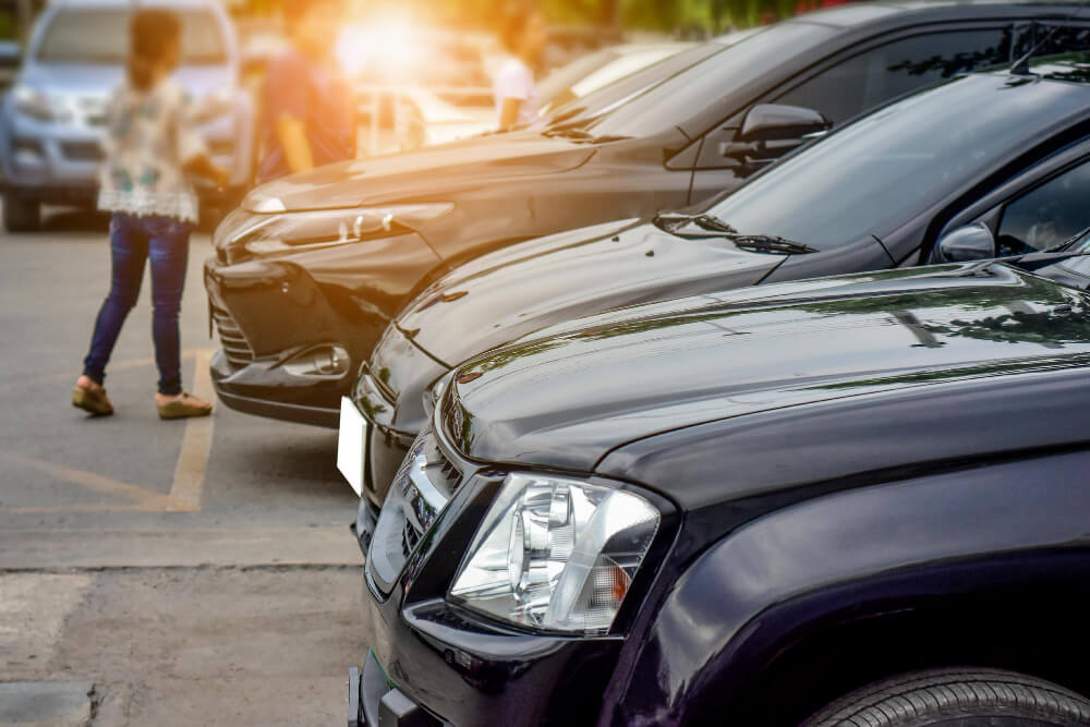 Mehrere parkende Autos auf einem Parkplatz, im Hintergrund gehen unscharfe Personen, Sonnenlicht fällt durch die Szenerie