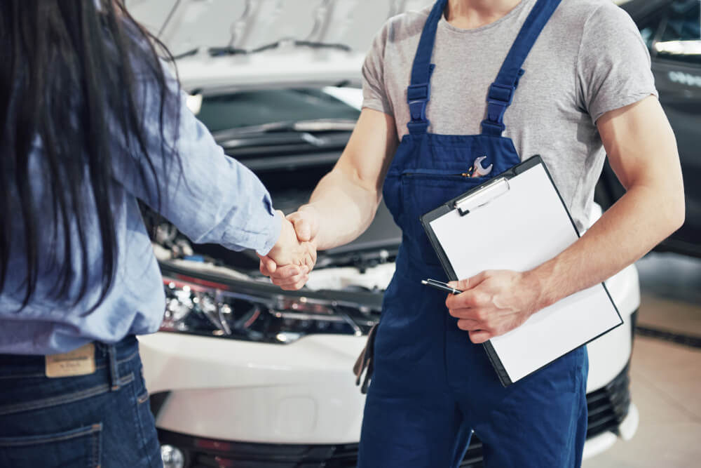 Ein Mechaniker in blauer Arbeitskleidung schüttelt nach einer Fahrzeugbewertung einem Kunden die Hand, neben einem Auto mit offener Motorhaube