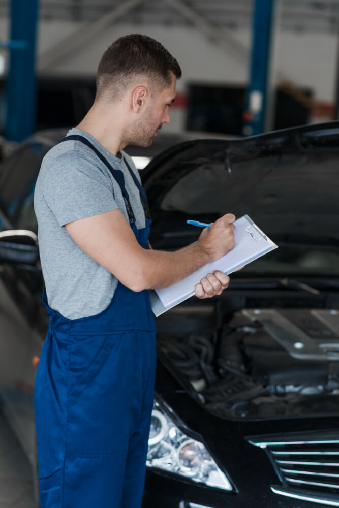Ein Mechaniker in blauer Arbeitskleidung prüft für ein Unfallgutachten den Motor eines Autos und macht Notizen auf einem Klemmbrett