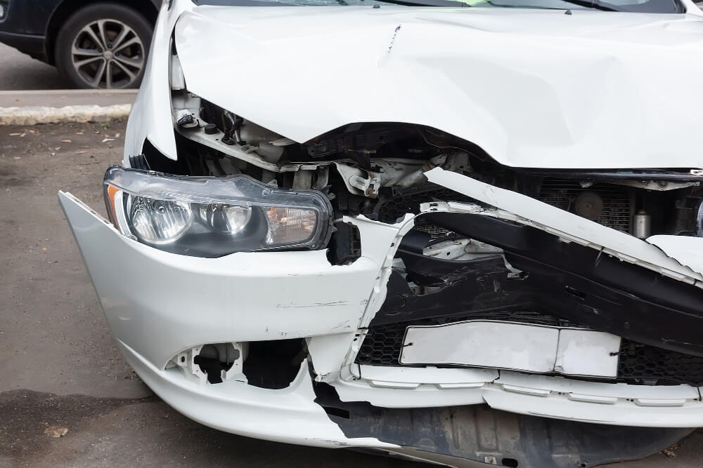 Ein weißes Auto mit massivem Frontschaden, zerbrochenem Scheinwerfer und eingedrückter Motorhaube nach einem Unfall