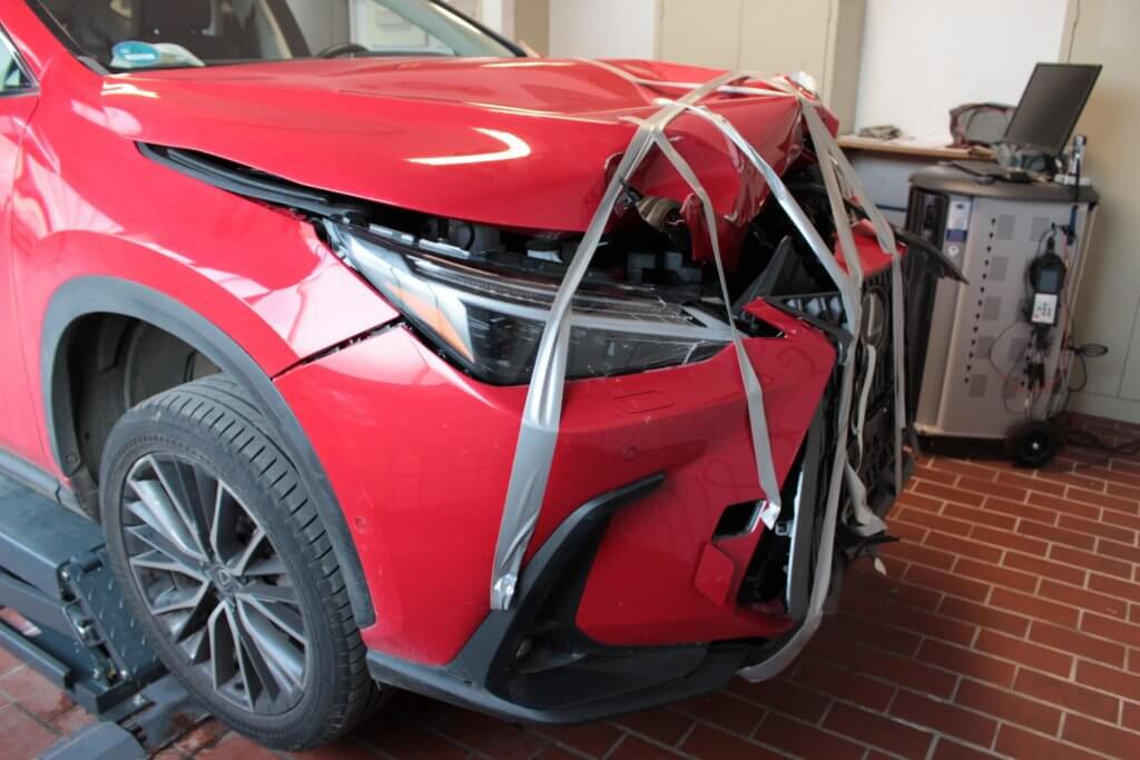Ein rotes Auto mit beschädigter Front, das mit Klebeband zusammengehalten wird, steht in einer Garage neben einem Computer