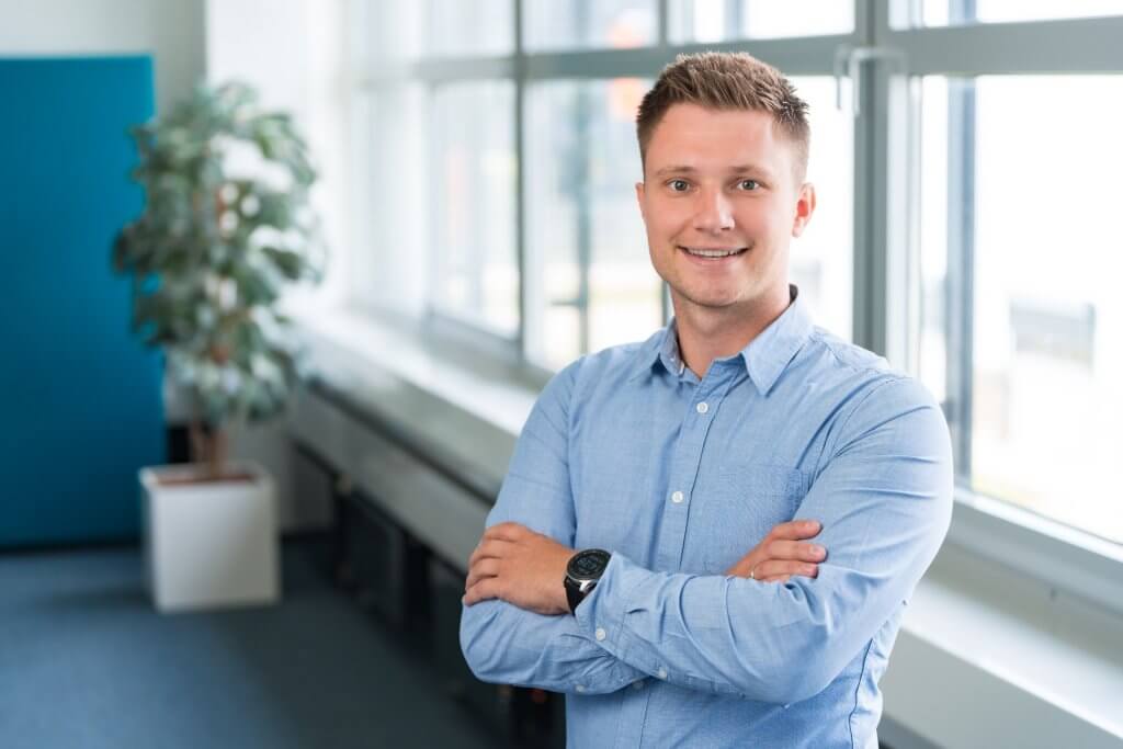 Ein lächelnder Mann im hellblauen Hemd steht mit verschränkten Armen in einem hellen Büro mit großen Fenstern und einer Topfpflanze