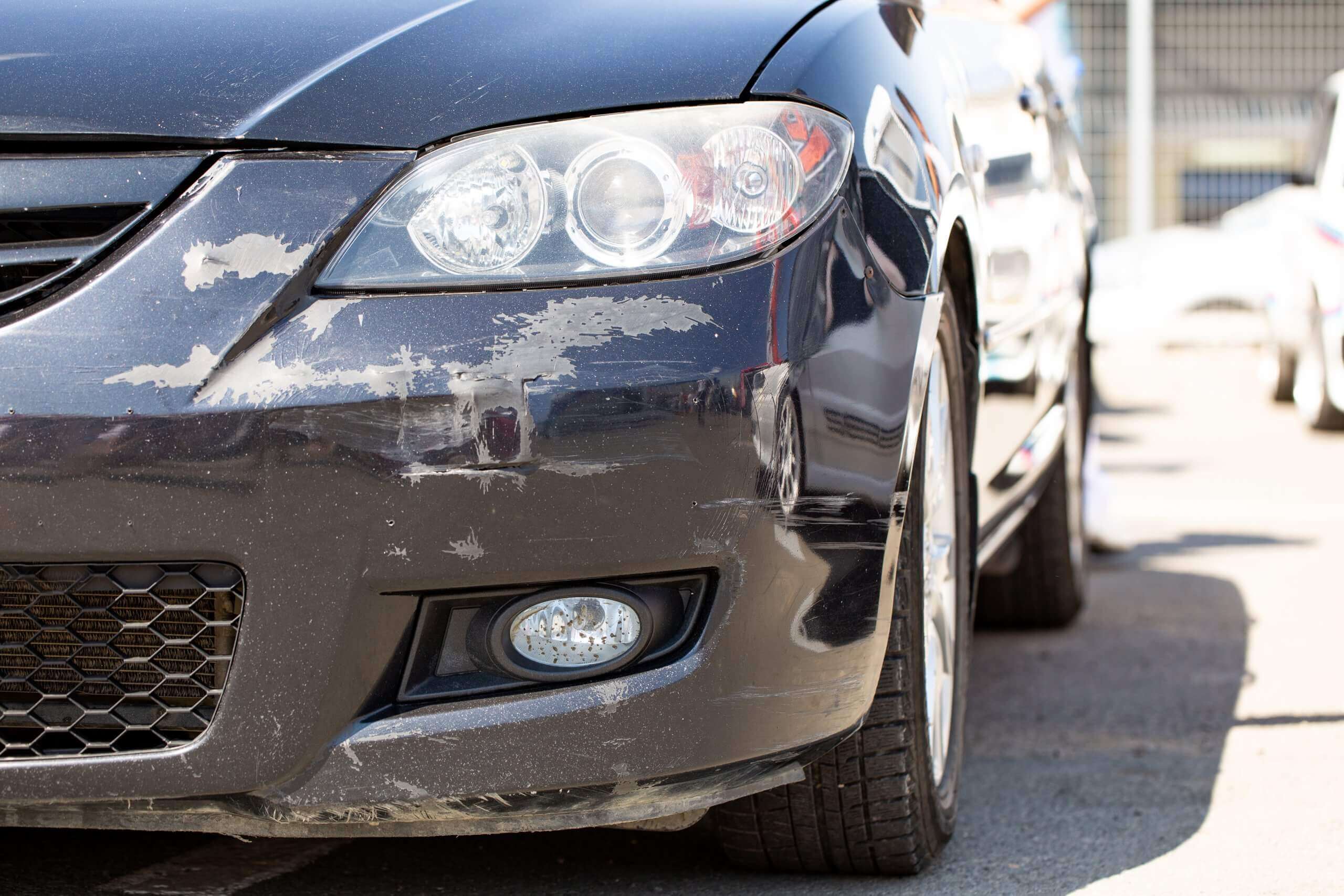 Vorderansicht eines schwarzen Autos mit Kratzern und Schrammen am Stoßfänger, draußen in der Sonne geparkt