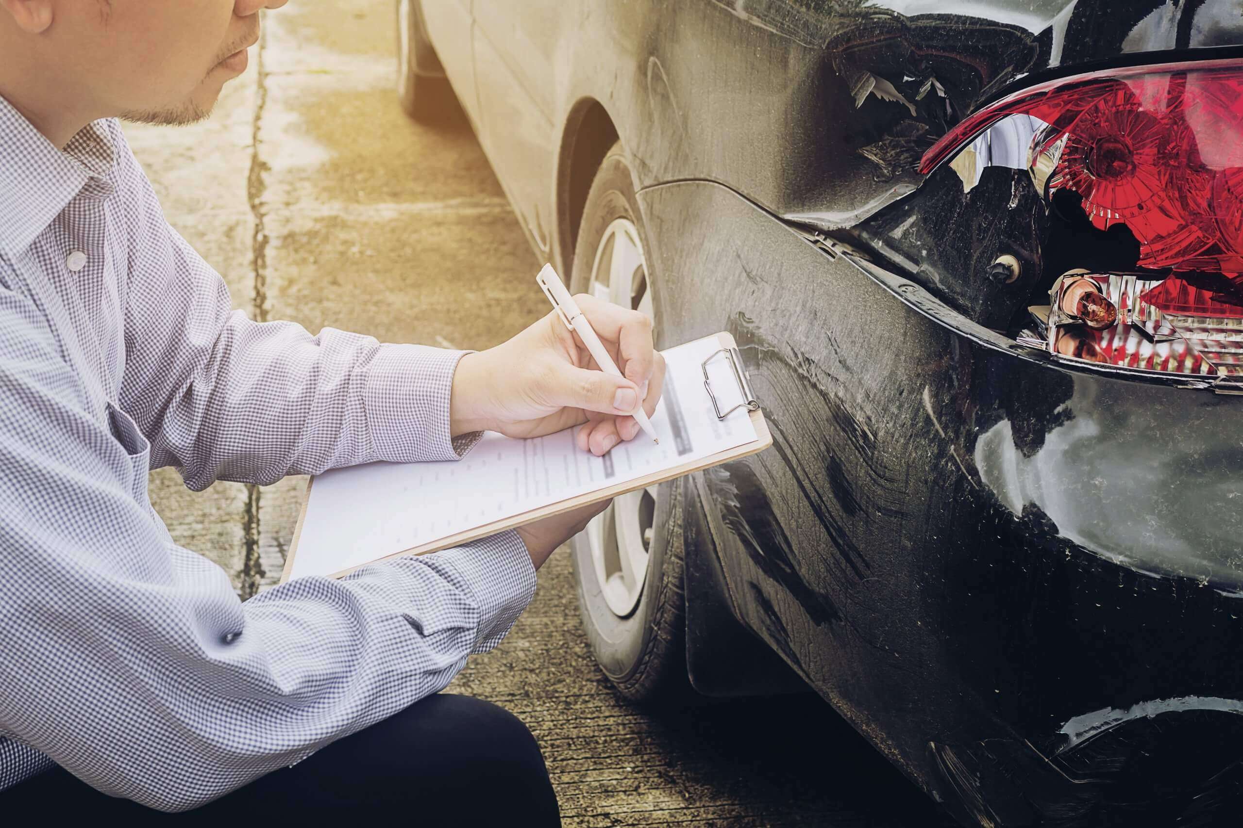 Ein Mann schreibt auf einem Klemmbrett neben einem schwarzen Auto mit Heckschaden und bewertet den Unfall als automatisch gespeicherten Entwurf