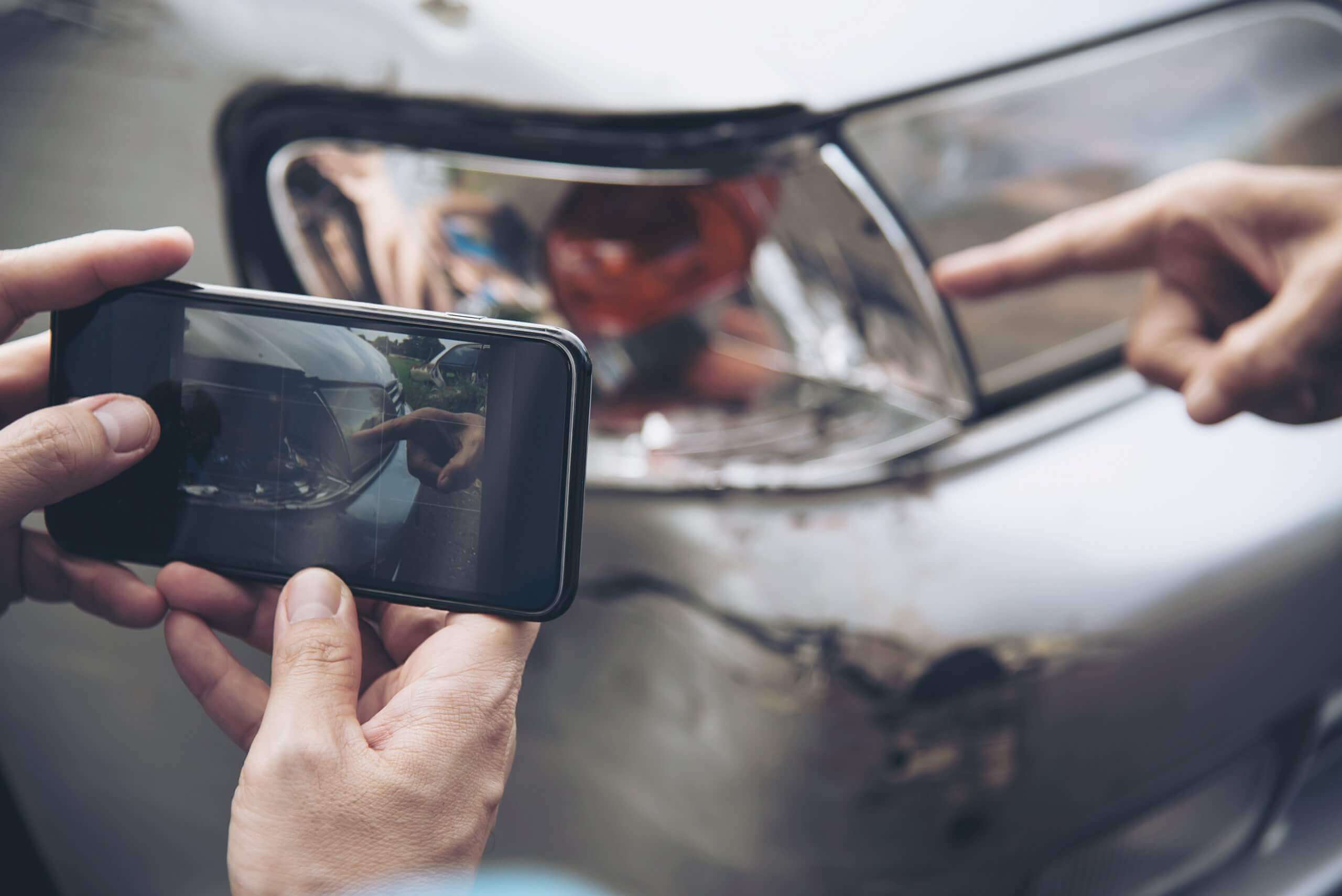 Eine Person fotografiert einen Unfallschaden am Auto, während eine andere Hand auf den verbeulten Scheinwerfer zeigt