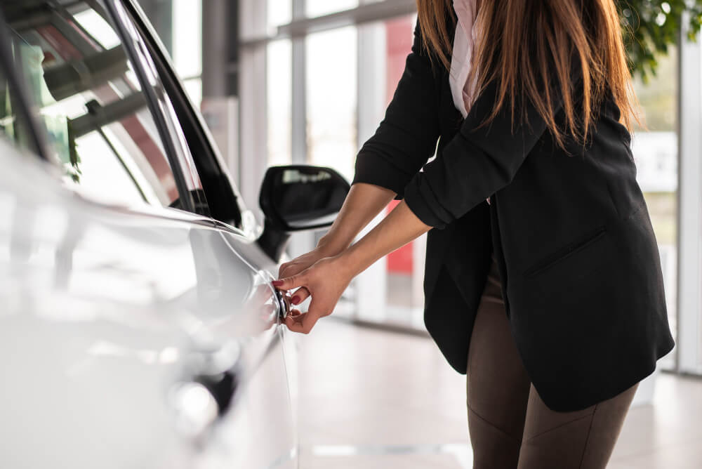 Eine Frau im Blazer öffnet die Tür eines weißen Autos in einem hellen Autohaus und schaut auf einen automatisch gespeicherten Entwurf