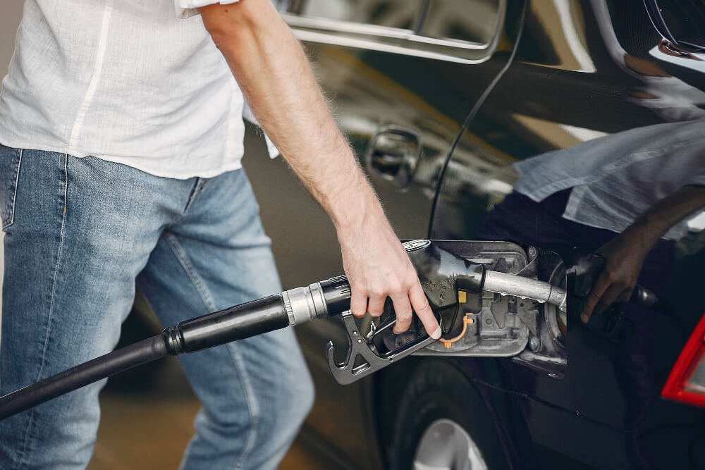 Eine Person in Jeans und weißem Hemd tankt ein schwarzes Auto an einer Tankstelle, neben einem automatisch gespeicherten Entwurf